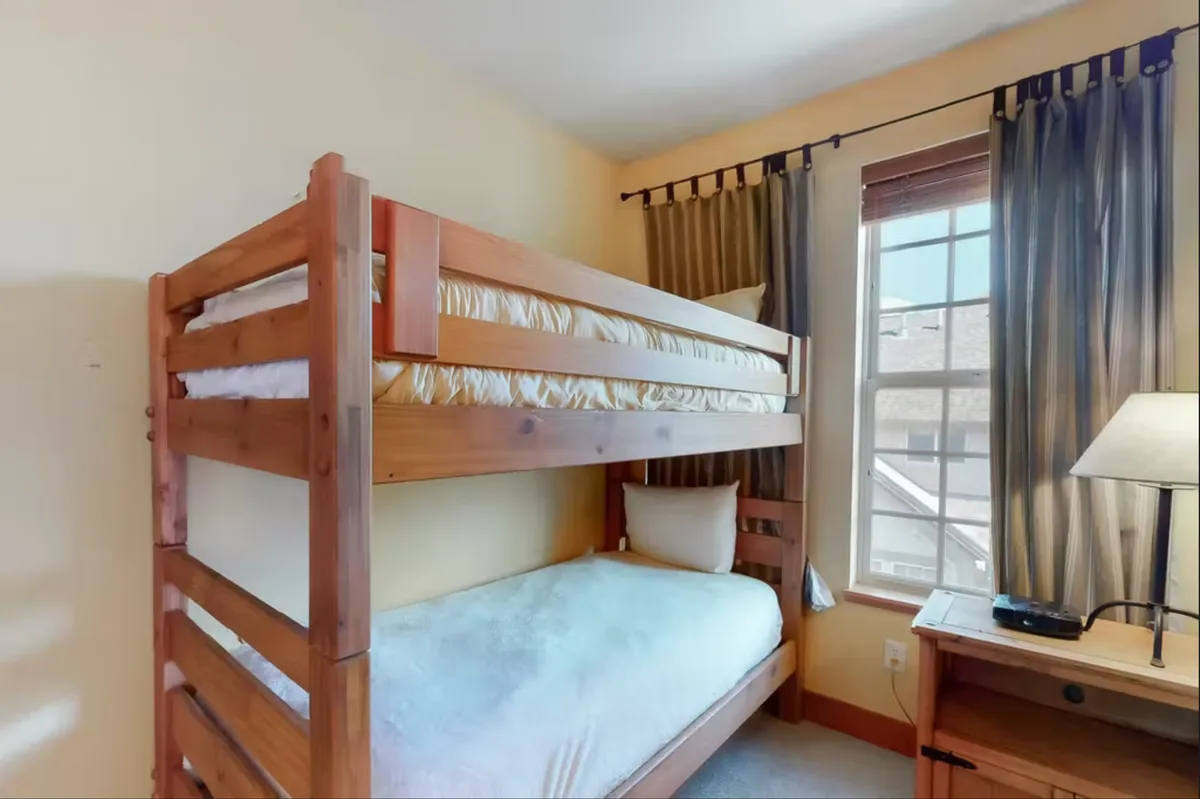 A cheerful bunkroom with a sturdy bunk bed, a window letting in soft daylight, a cozy lamp, and a convenient nightstand. The space feels playful and welcoming, ideal for children or young-at-heart guests to enjoy sleepovers, quiet reading, or games.