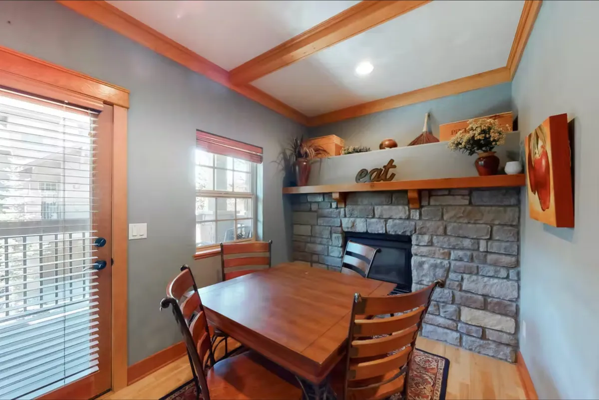 A cozy dining area centered around a spacious dining table, set near a stone fireplace and window. The decor is intimate and welcoming, inviting family and friends to gather for meals, coffee chats, or holiday celebrations.