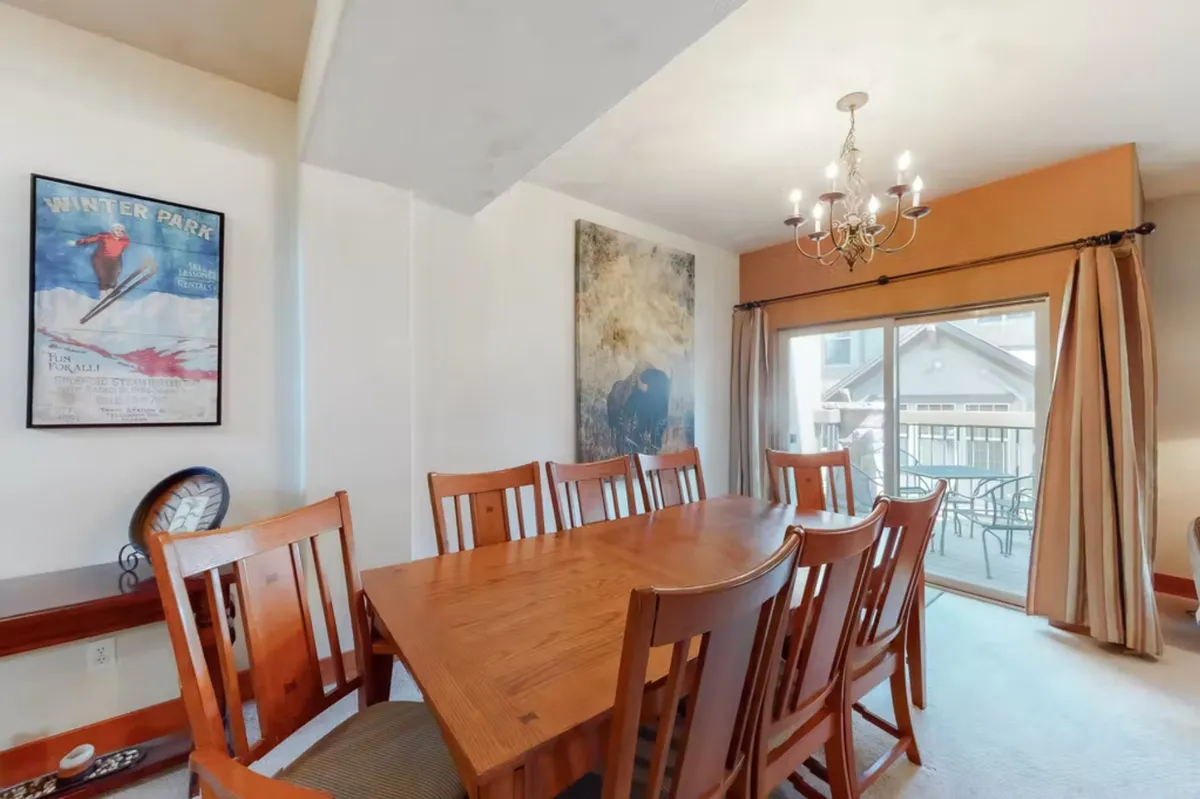 A view of a spacious, open dining area with a large wooden table, an elegant chandelier, decorative artwork, and a sliding glass door leading out to the balcony. The inviting table is set for family dinners, with plenty of seating to bring groups together.