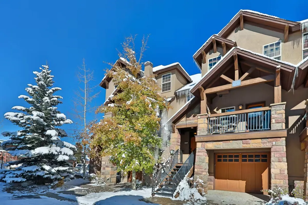 A picturesque scene featuring a building with a stone facade and wooden balcony, set against a landscape of snow-covered trees. The area around the building is blanketed in fresh snow, evoking a sense of winter adventure and outdoor activity. The cozy appearance suggests the perfect home base for winter sports or après-ski relaxation.