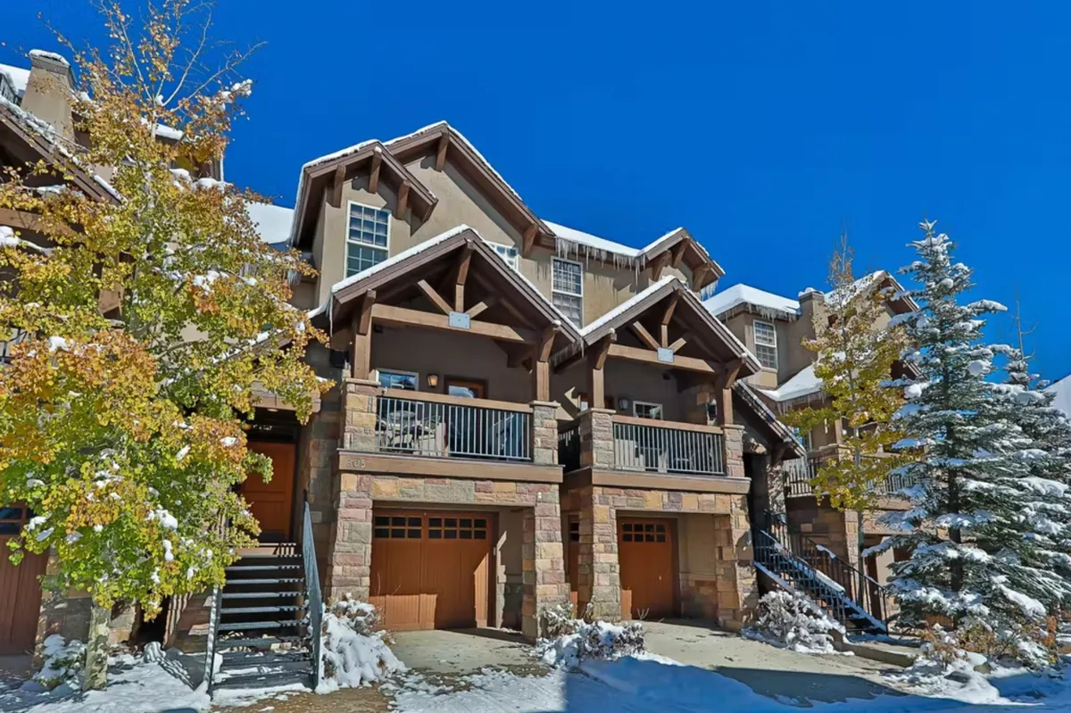 The image showcases the elegant exterior of a luxury mountain villa, with warm wooden balconies and a striking stone facade nestled among colorful autumn trees. Snow-dusted rooftops hint at the winter wonderland beyond, while sweeping balconies invite guests to relax and soak in the panoramic alpine views.