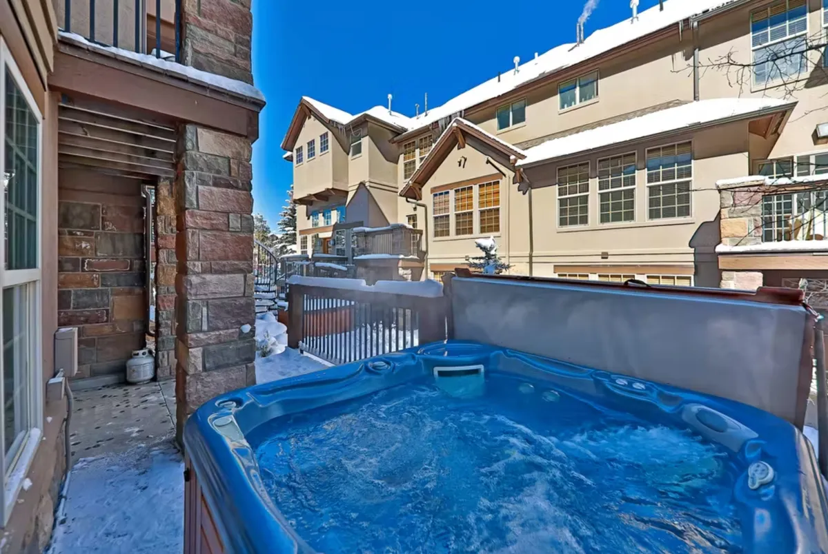 An exterior view of the property features a bubbling hot tub nestled in a snow-covered setting. The inviting hot tub sits on a deck with the building's architecture in the background, while people are seen relaxing and enjoying the warmth after a day outdoors. Snow-dusted surroundings enhance the cozy, rejuvenating mood, and the hot tub is clearly the focal point.