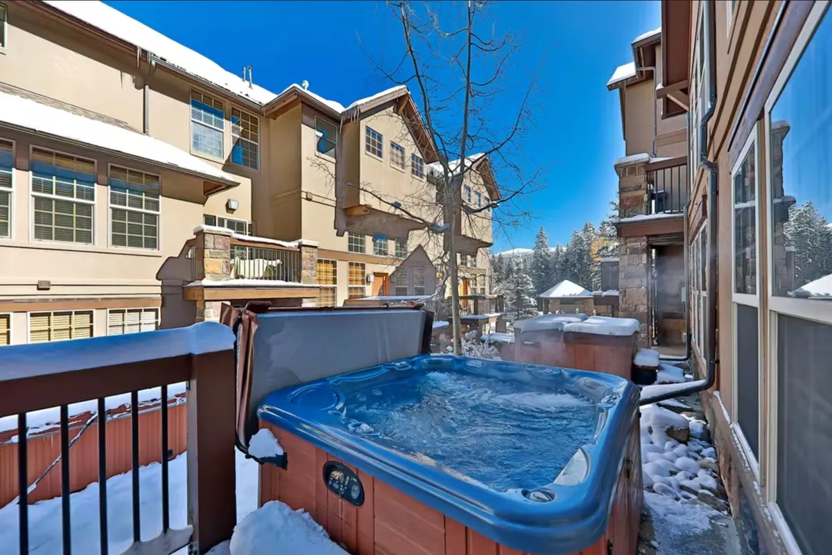 This exterior photo features a steaming hot tub set on a wooden deck, surrounded by snow and neighboring buildings. The scene invites relaxation after a day exploring Winter Park, with the juxtaposition of warm water and brisk mountain air.