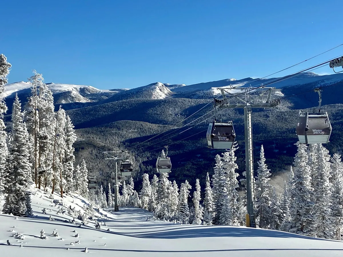 A breathtaking exterior scene of a ski lift gliding above snow-covered trees and a stunning mountain range under a clear blue sky. Skiers and snowboarders can be spotted below, with the scene radiating both tranquility and exhilarating opportunity.