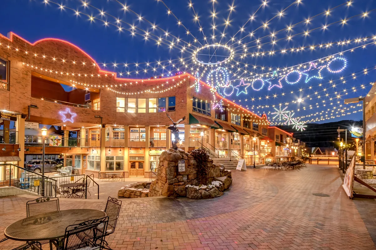 A lively outdoor plaza illuminated by string lights, with decorative star motifs, a stone fountain, and ample outdoor seating. People can be seen strolling, dining, and enjoying a festive atmosphere. The scene suggests a vibrant village or mountain resort base, bustling with activities and social gatherings in any season.