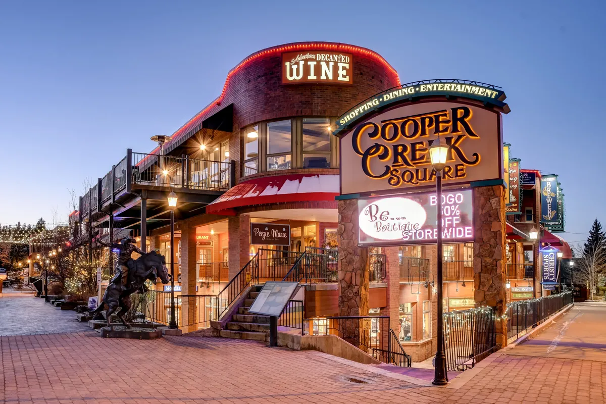 This image captures the festive atmosphere of Cooper Creek Square in downtown Winter Park, featuring the prominent square sign, wine shop signage, inviting outdoor seating areas, and twinkling decorative lighting. The environment is lively and welcoming, with visual cues suggesting shopping, dining, and entertainment options just steps away.