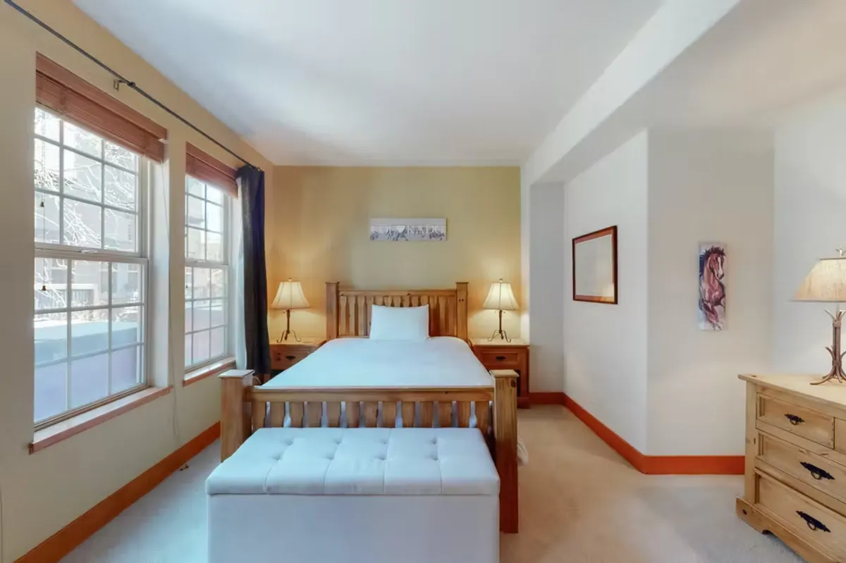 A serene bedroom featuring a plush bed with premium bedding, flanked by stylish nightstands and lamps. A bench sits at the foot of the bed, while tasteful artwork adorns the wall. Sunlight filters gently through a nearby window, creating a restful, inviting atmosphere.